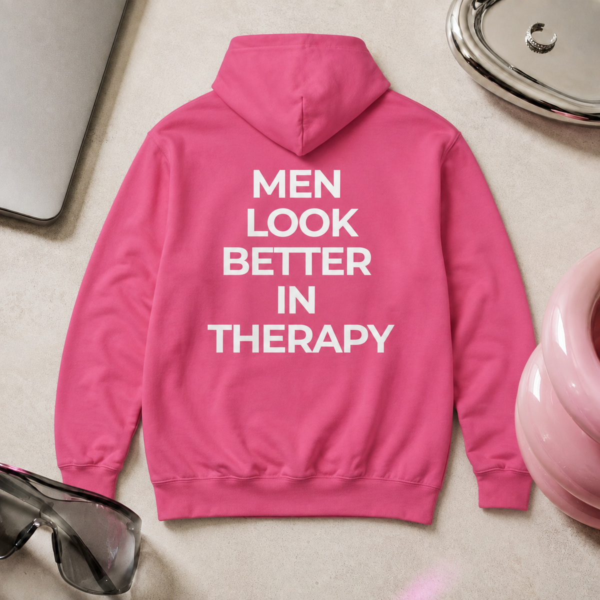 Bright pink unisex hoodie with a white “Men Look Better in Therapy” statement print across the back.
