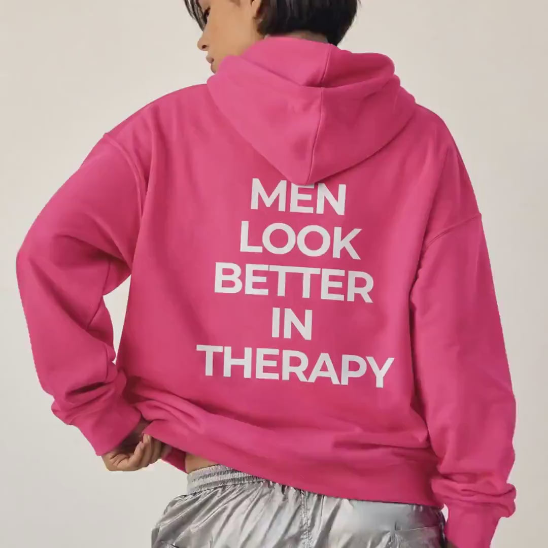 Bright pink unisex hoodie with a white “Men Look Better in Therapy” statement print across the back.
